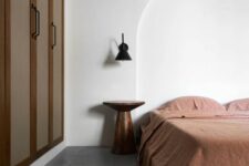 a minimal earthy bedroom with a built-in wardrobe, a bed with rust bedding, a metal nightstand and a black sconce