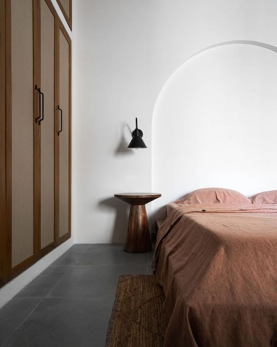 a minimal earthy bedroom with a built-in wardrobe, a bed with rust bedding, a metal nightstand and a black sconce