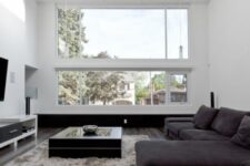 a minimalist black and white living room with double-height ceilings and a skylight, a black sectional, a TV and a low coffee table on a grey rug
