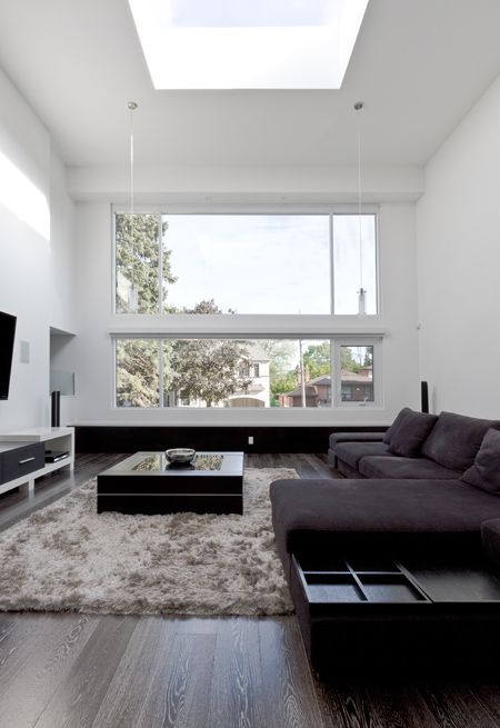 a minimalist black and white living room with double height ceilings and a skylight, a black sectional, a TV and a low coffee table on a grey rug