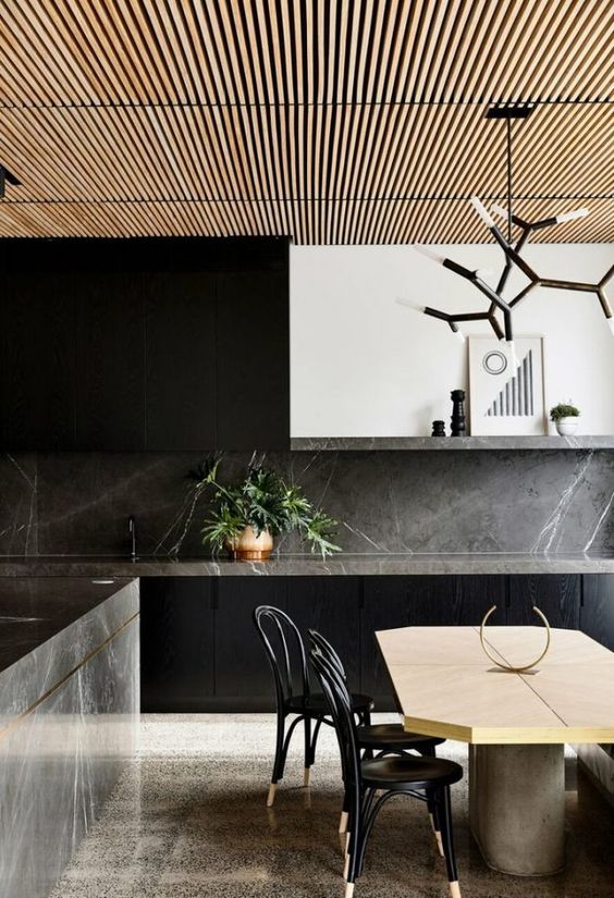 a minimalist black kitchen with flat panel cabinets, grey soapstone countertops and a backsplash, a white hood and a soapstone kitchen island