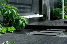 a minimalist black outdoor space with weathered oak millboard decking, a waterfall, a wall-mounted bench and some greenery plus a pond
