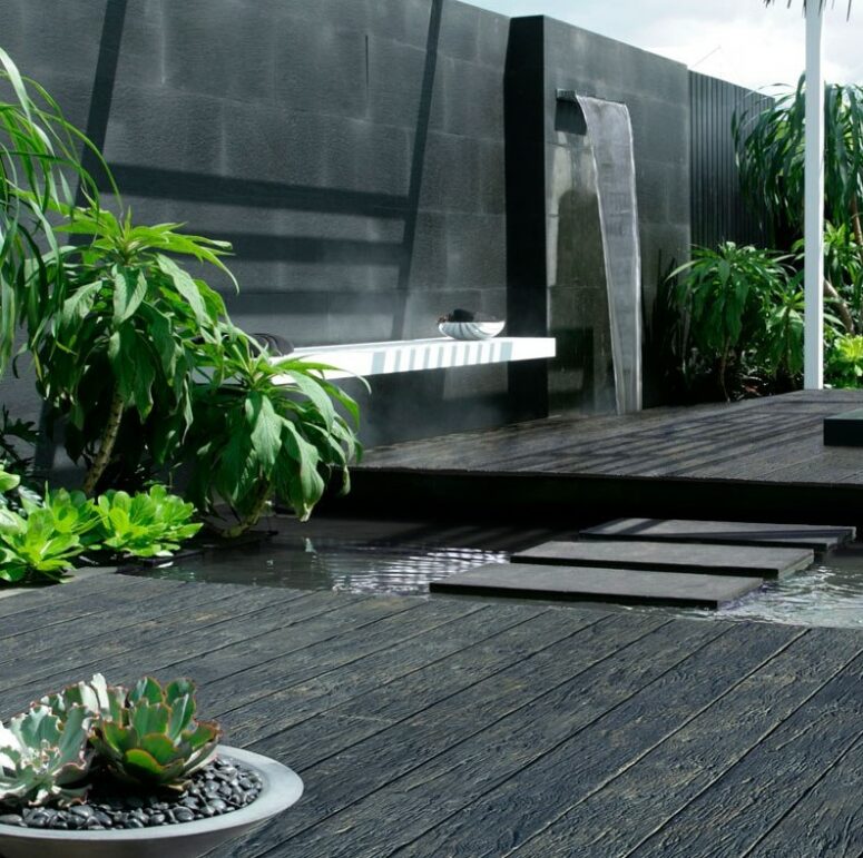 a minimalist black outdoor space with weathered oak millboard decking, a waterfall, a wall-mounted bench and some greenery plus a pond