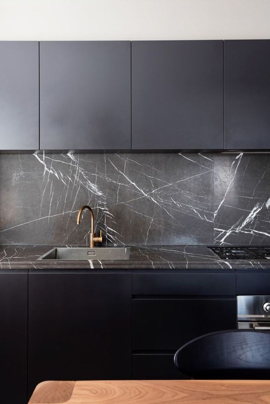 a minimalist midnight blue kitchen with sleek cabinets, a black soapstone backsplash and countertops plus brass fixtures