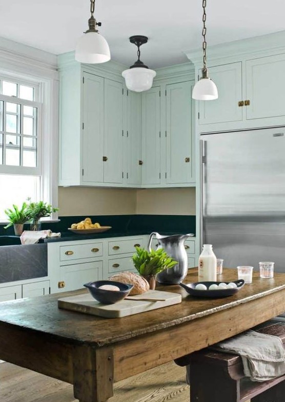 a mint green farmhouse kitchen black granite countertops and a backsplash, brass handles, a stained rough wood table and lamps on chain