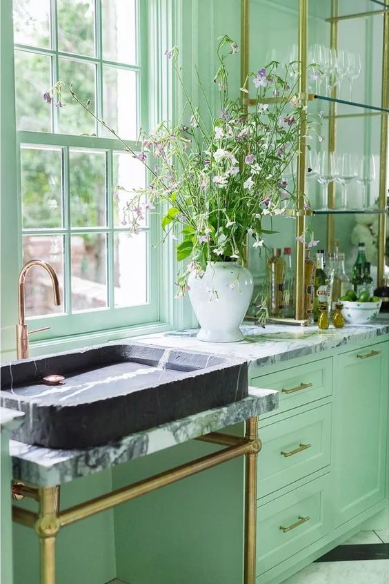 a mint grene kitchen with shaker cabinets, a glass shelving unit, a marble sink and a sink stand and some brass touches