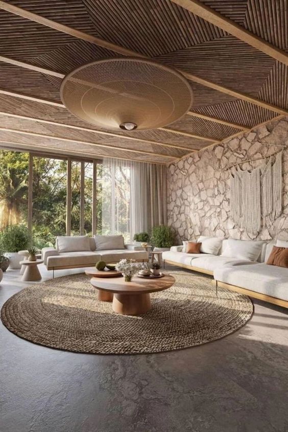 a modern earthy living room with a tropical feel, a stone accent wall, a wooden ceiling, a large rug, white sofas and chairs, potted greenery