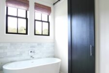 a modern farmhouse bathroom with black frame double-hung windows, a marble tile backsplash and an oval tub