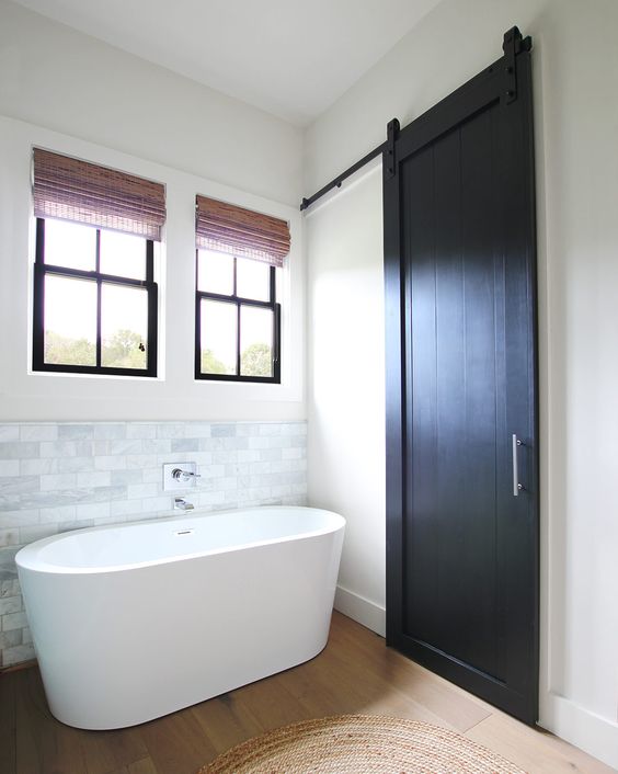 a modern farmhouse bathroom with black frame double hung windows, a marble tile backsplash and an oval tub
