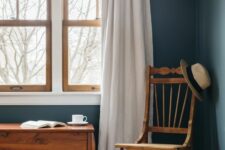 a modern farmhouse space with navy walls, a stained chair and chest and echoing stained double-hung windows and white curtains
