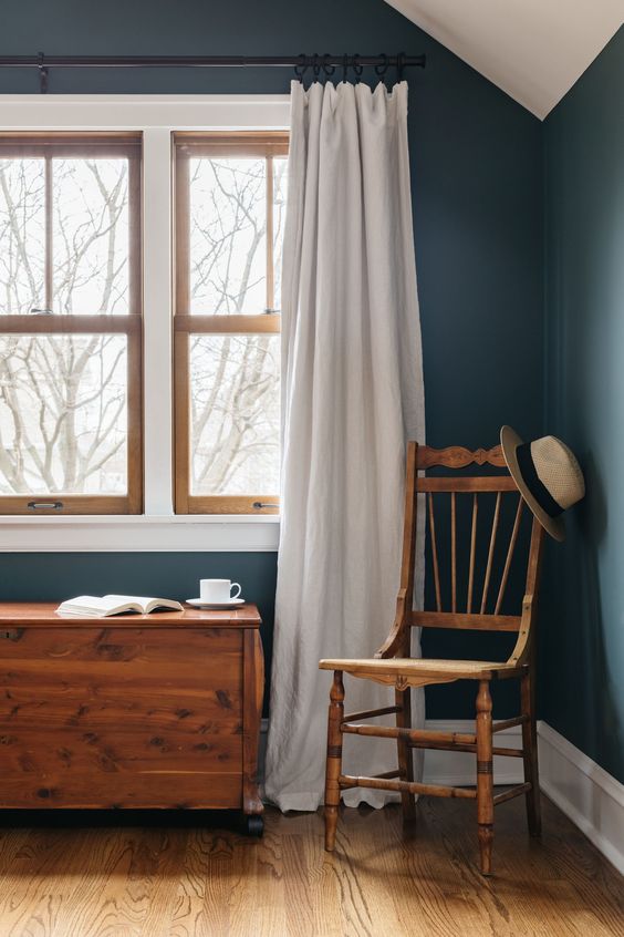 a modern farmhouse space with navy walls, a stained chair and chest and echoing stained double-hung windows and white curtains