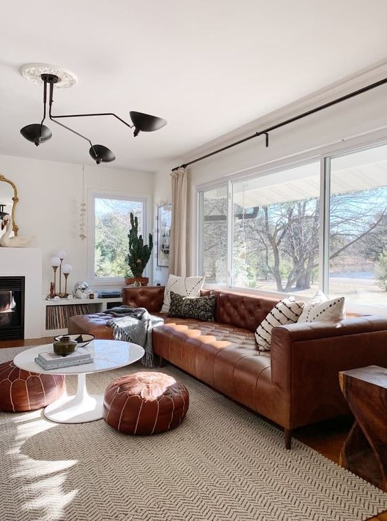 a modern neutral living room with a fireplace, a brown leather Chesterfield sofa, matching ottomans, a black chandelier and a glazed wall