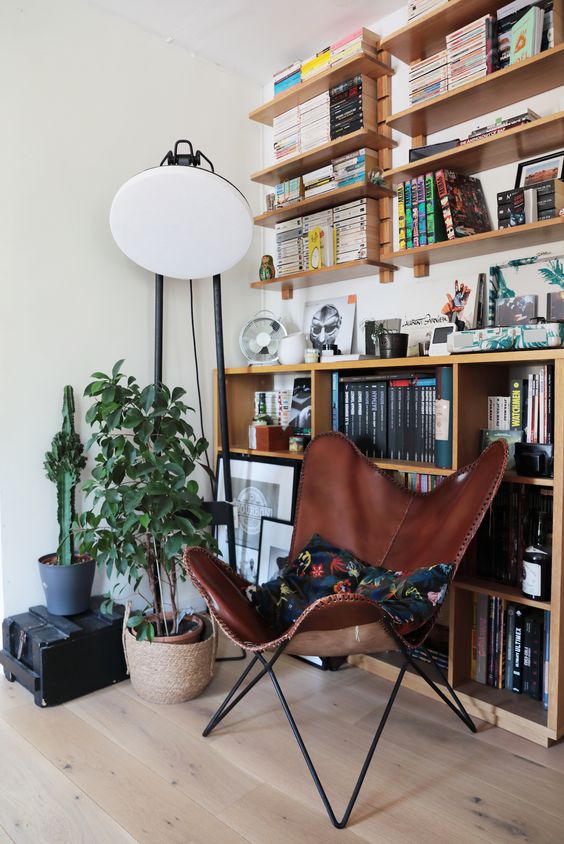 a modern nook with bookshelves and a large shelving unit, a brown leather butterfly chair, potted plants and a black lamp