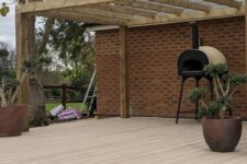 a modern outdoor cooking space clad with limed oak decking, with a wooden frame over the space, a hearth and some potted plants