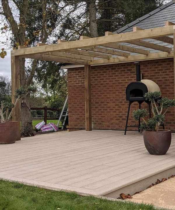 a modern outdoor cooking space clad with limed oak decking, with a wooden frame over the space, a hearth and some potted plants