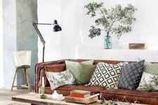 a modern rustic living room with a brown leather sofa, a wooden bench as a coffee table, printed pillows, greeneyr and a black lamp