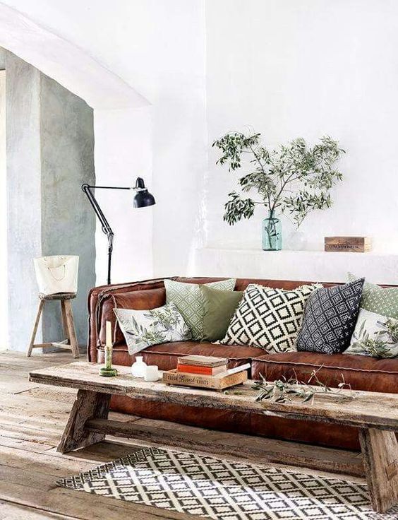 a modern rustic living room with a brown leather sofa, a wooden bench as a coffee table, printed pillows, greeneyr and a black lamp