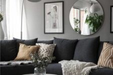 a monochromatic living room with a grey wall, a black sectional, printed pillows, artworks and a round mirror, a coffee table and a pouf