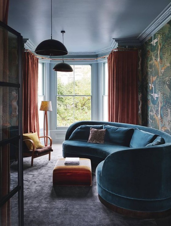 a moody and bold living room with a curved blue velvet sofa, orange textiles and moody bird print wallpaper on one wall