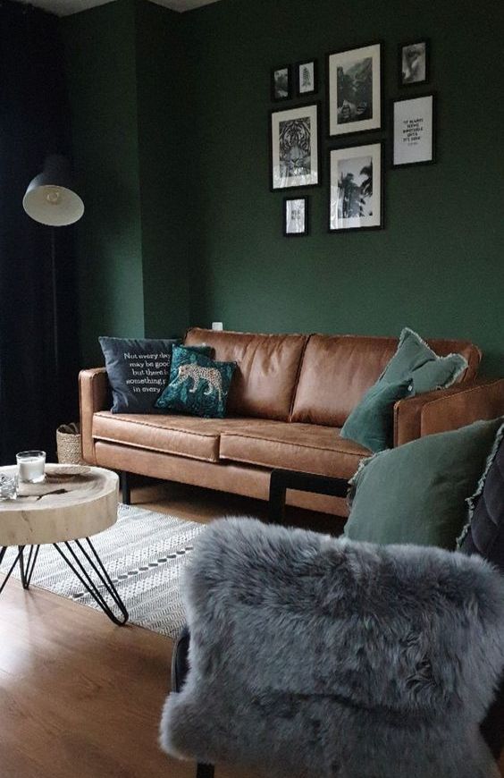 a moody living room with dark green walls, a brown leather sofa, a gallery wall, a grey chair and a tree slice coffee table