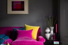 a moody living room with grey walls, a hot pink sofa, a yellow tufted ottoman, a printed rug, chic vases and candles