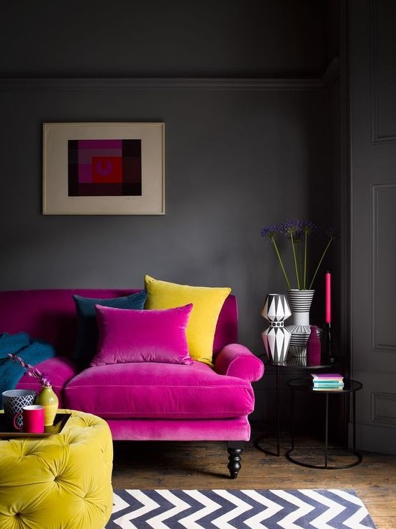 a moody living room with grey walls, a hot pink sofa, a yellow tufted ottoman, a printed rug, chic vases and candles