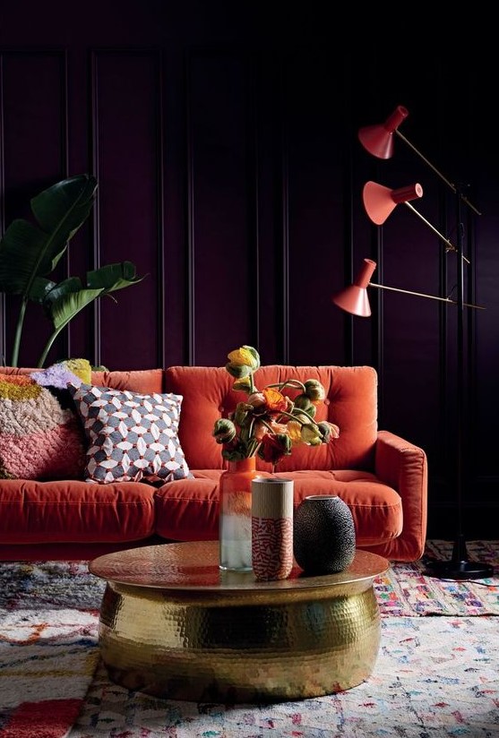 a moody living room with purple paneled walls, an orange sofa, orange floor lamps, a gold hammered table and statement plants