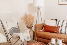 a neutral boho space with a tan leather sofa and a rust-colored ottoman, striped textiles, pampas grass and a wooden table