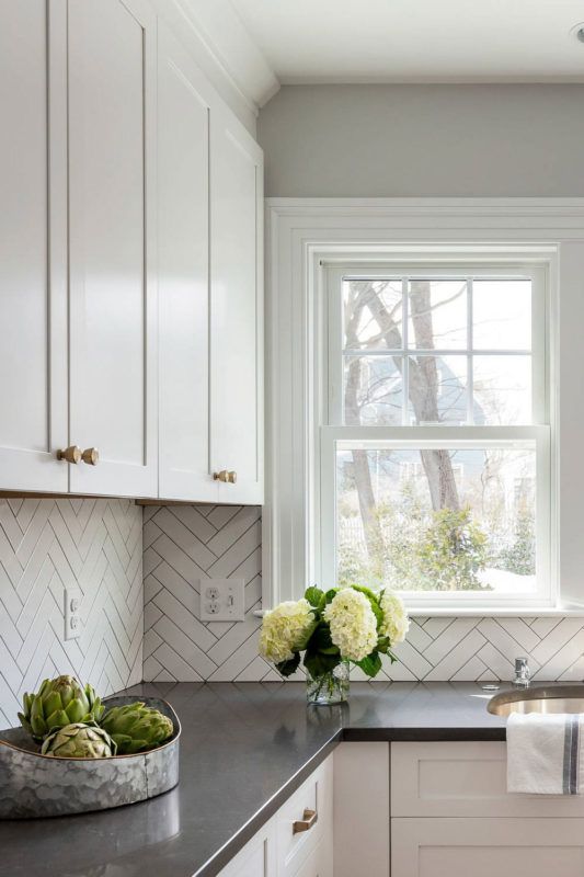 a neutral farmhouse kitchen with shaker cabinets, black countertops, a white herringbone tile backsplash and a small double hung window