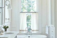 a neutral vintage bathroom with trim, an oval tub, a double-hung window, curtains, a double sink on a metal stand