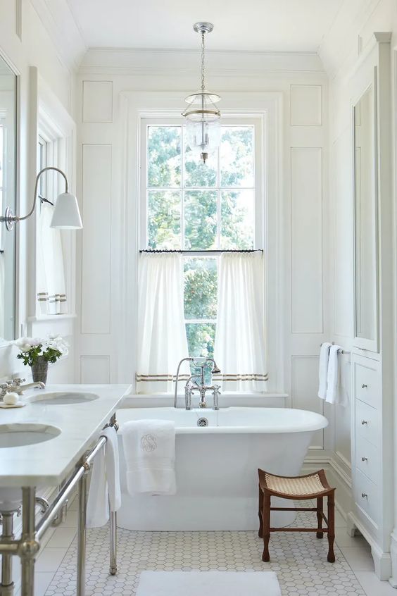 a neutral vintage bathroom with trim, an oval tub, a double-hung window, curtains, a double sink on a metal stand