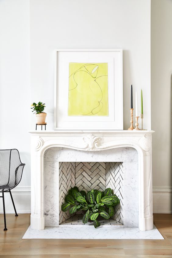 a non-working French fireplace with a chic ornated mantel, a bold artwork and a potted plant inside it