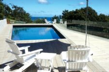 a pool space with smoked oak decking, white folding furniture and a table, an umbrella and greenery around