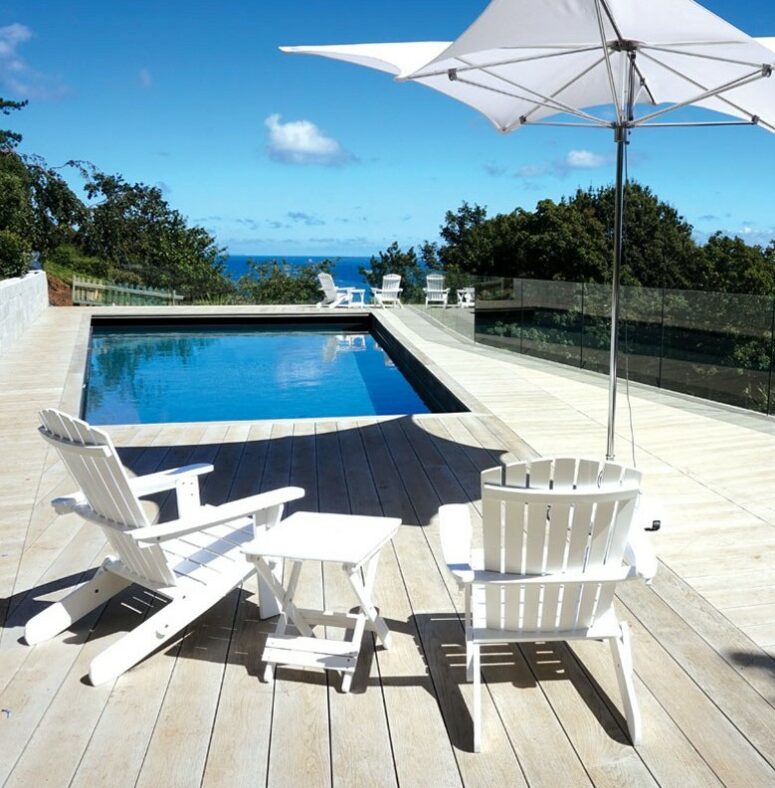 a pool space with smoked oak decking, white folding furniture and a table, an umbrella and greenery around