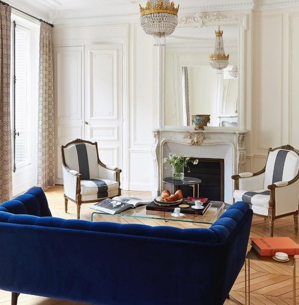 a refined French style living room with a non-working French fireplace, a bold blue sofa, striped vintage chairs, a chic crystal chandelier and a clear acrylic table