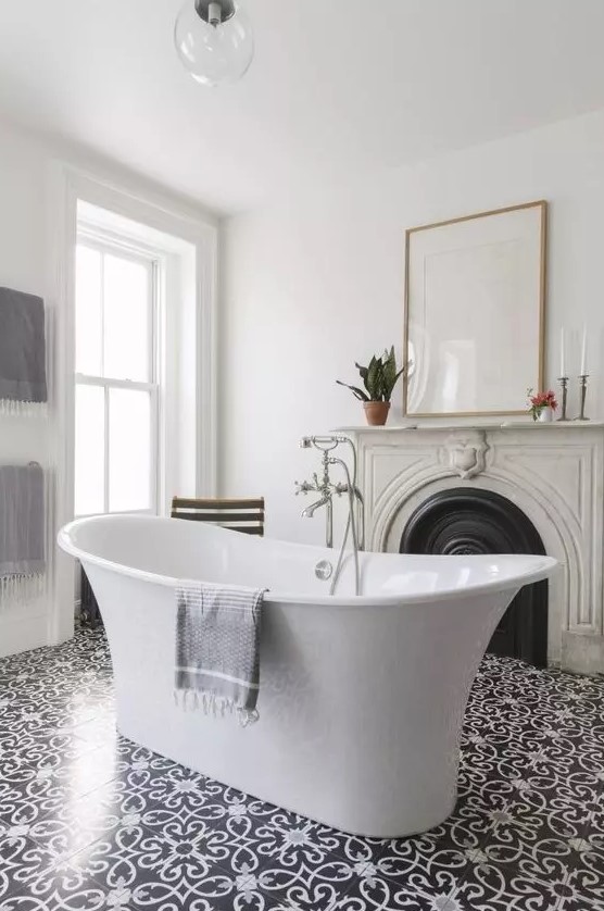a refined and chic bathroom with a vintage French fireplace with a stone mantel, a free-standing tub, some art and beautiful tiles on the floor