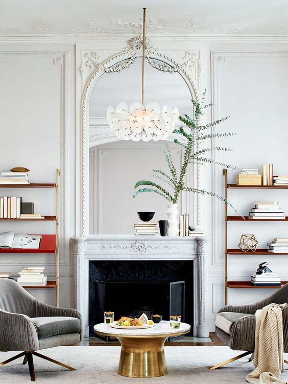 a refined living room with a French fireplace and an arched mirror in an ornated frame, bookshelves, a round coffee table and grey chairs