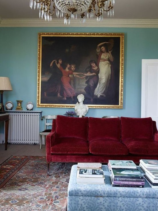 a refined living room with blue walls, a deep red sofa, a floral ottoman, a bold vintage artwork and a crystal chandelier