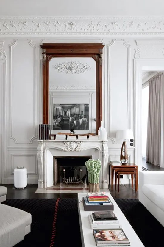 a refined living room with molding and an ornated French fireplace, white seating furniture, a coffee table, a couple of oversized mirrors