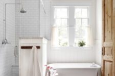 a rustic bathroom clad with subway tiles, with a wooden floor, a clawfoot tub and shower space plus a double-hung window with curtains