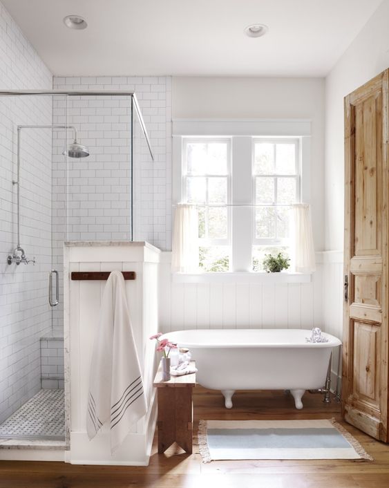 a rustic bathroom clad with subway tiles, with a wooden floor, a clawfoot tub and shower space plus a double hung window with curtains