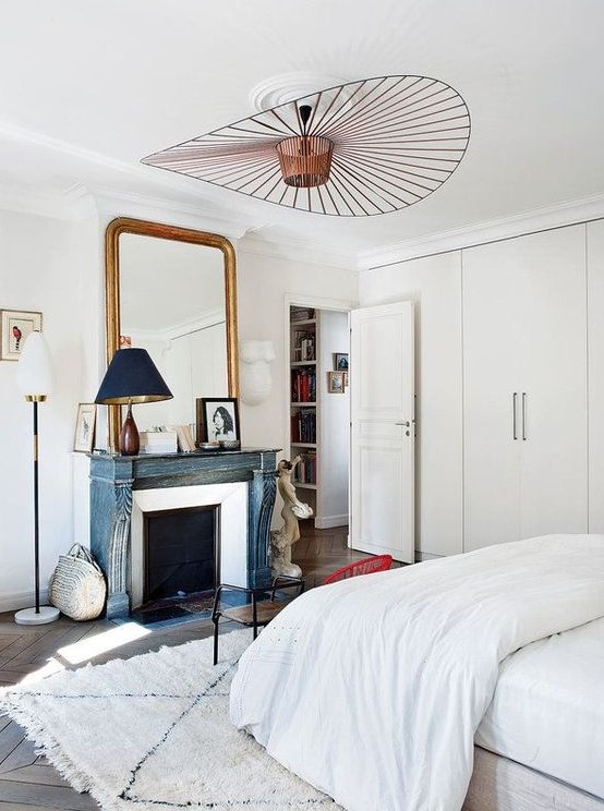 a serene Parisian bedroom with built-in wardrobes, a French fireplace with a blue mantel, a large mirror in a gilded frame and a bed with neutral bedding
