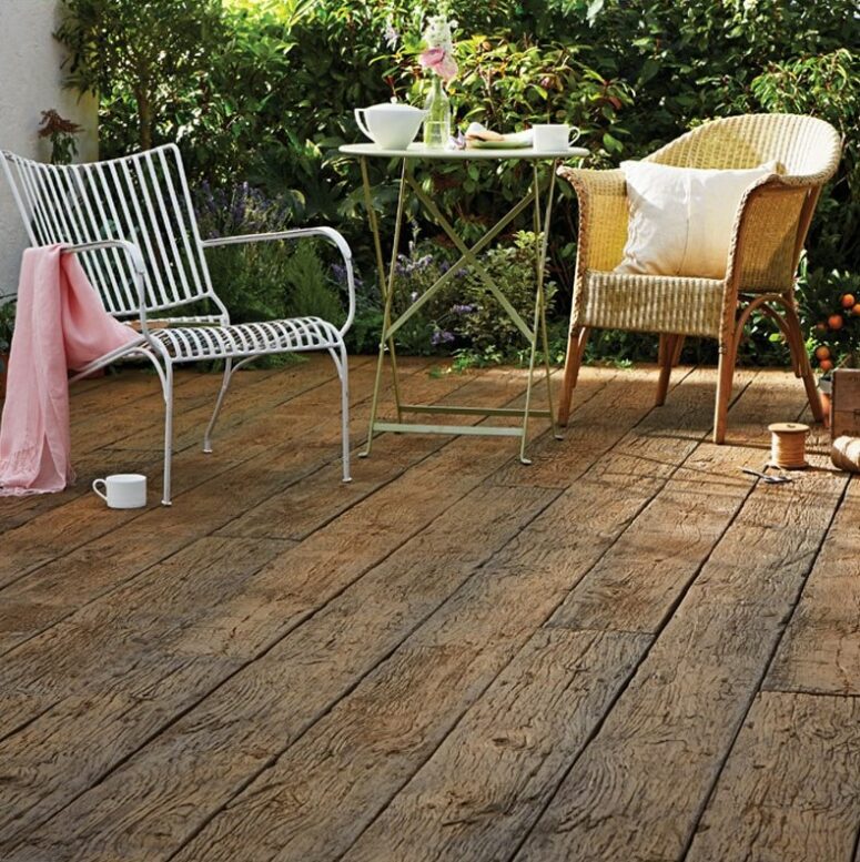 a small and cute outdoor space done with weathered oak, with a metal coffee table anda woven and a metal chair plus greenery in the backdrop