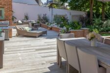 a smoked oak millboard deck with neutral upholstered furniture, a daybed for two, an outdoor kitchen and lots of greenery and blooms