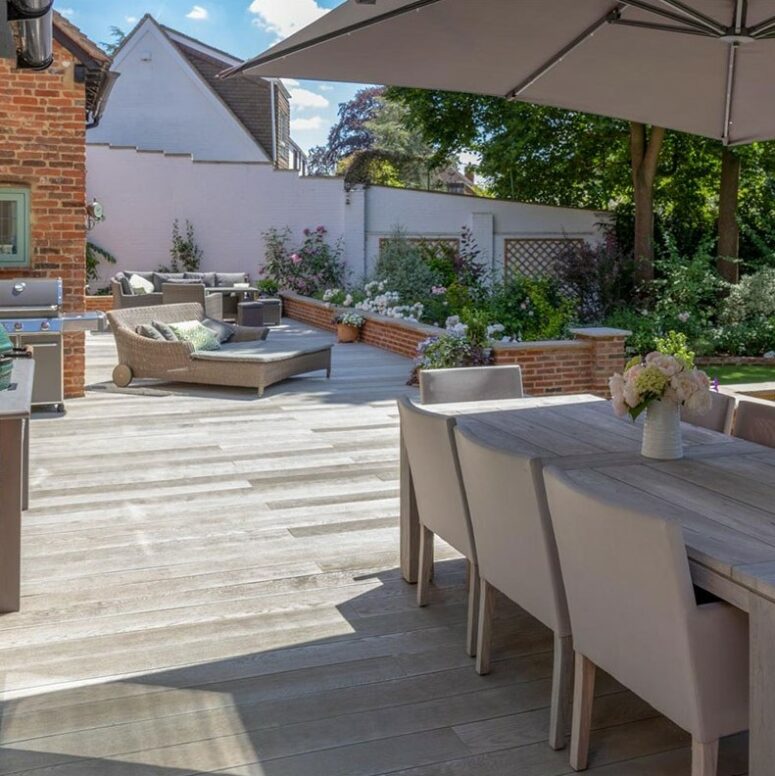 a smoked oak millboard deck with neutral upholstered furniture, a daybed for two, an outdoor kitchen and lots of greenery and blooms