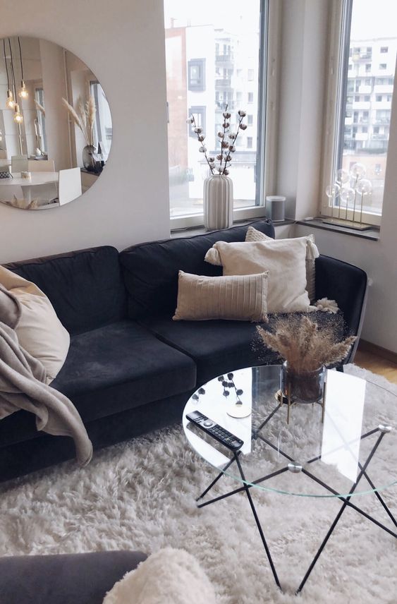 a soft and cozy living room with a black sofa, a clear glass hairpin leg table, neutral pillows and a round mirror