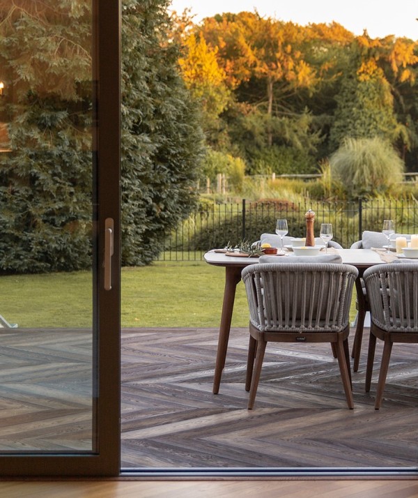 a sophisitcated modern terrace clad with antique oak composite decking, with a modern dining table and an upholstered bench plus a beautiful garden next to the deck