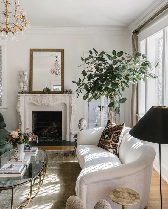 a sophisiticated neutral space with a French fireplace, a white curved sofa, an oval coffee table, a potted plant