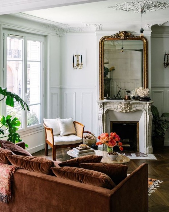a sophisticated French chic living room with a French fireplace, a large mirror in an ornated frame, a neutral chair, a rust-colored sofa with pillows and potted plants