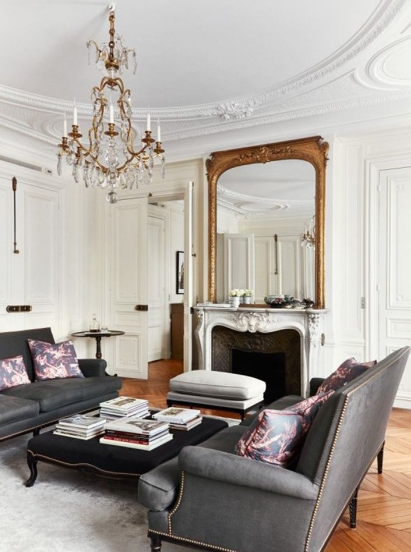 a sophisticated French style living room with a French fireplace, graphite grey sofas, an elegant black ottoman and a crystal chandelier